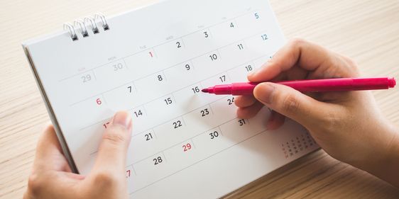 Terminplanung im Kalender Eine Person markiert mit einem pinkfarbenen Stift ein Datum in einem Tischkalender. Der Kalender zeigt einen Monatsüberblick mit Zahlen von 1 bis 31, wobei die Sonntage in rot markiert sind. Die Person hält den Kalender mit einer Hand fest, während sie mit der anderen Hand den Stift führt.